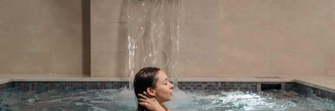 Adult woman relaxing in an indoor swimming pool near a cascading waterfall.